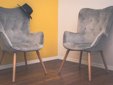 Two contemporary gray armchairs in a minimalist interior with a yellow accent wall and wooden flooring.