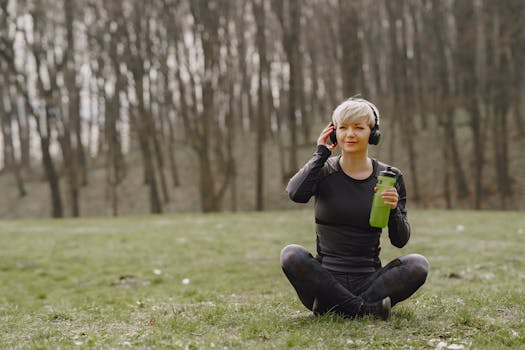 Woman sitting cross-legged in forest park with headphones and water bottle, enjoying a peaceful moment.