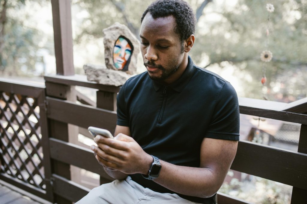 pexels-photo-6669804-6669804 African American man sitting on a deck, focused on his smartphone outdoors.